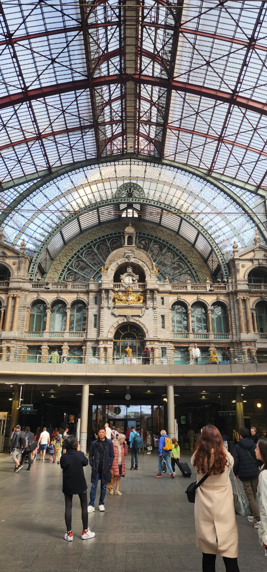 Antwerp train station exterior with a grand glass ceiling, intricate architecture, and travelers walking through the entrance.