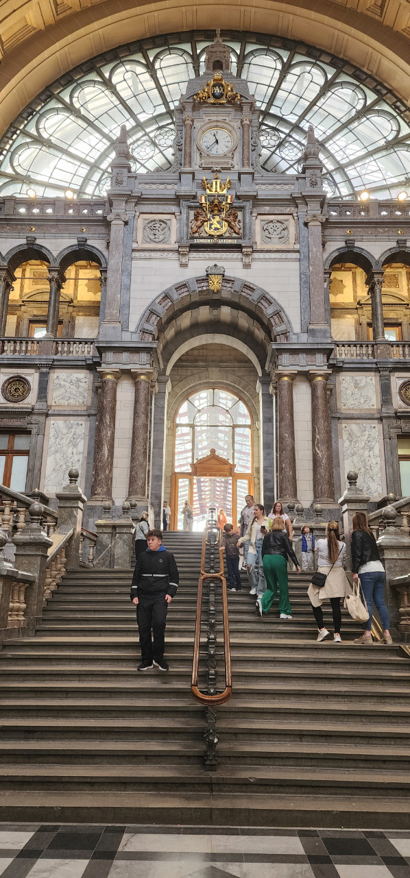 The interior of Antwerp train station, featuring a grand staircase, marble detailing, and an ornate clock with gold accents.