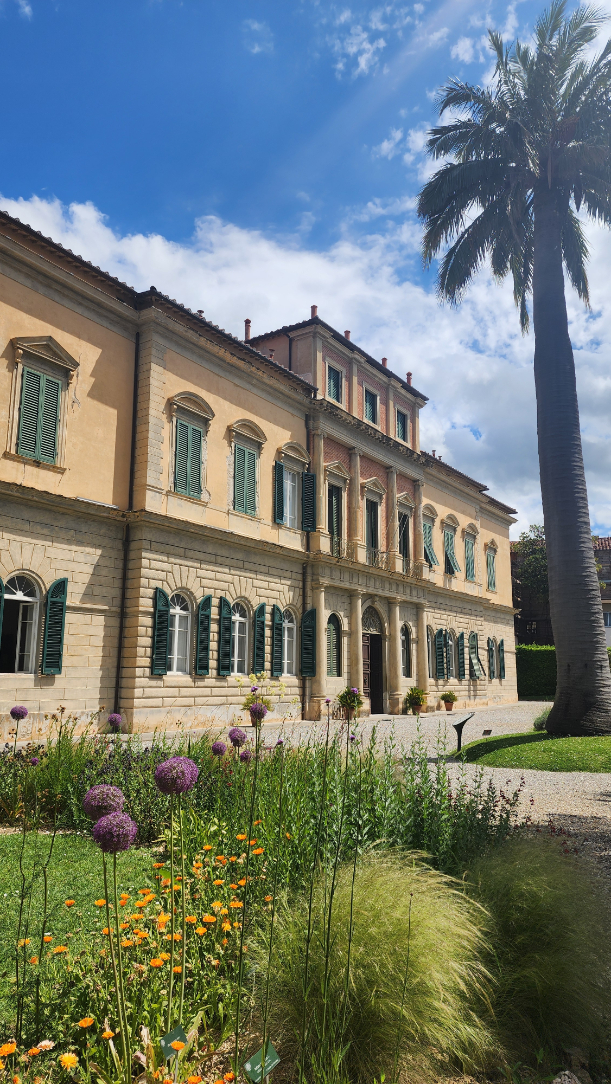 The botanical gardens of Pisa, showcasing lush greenery, blooming purple alliums, and an elegant historic villa with green shutters.