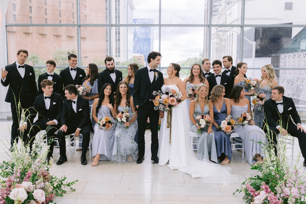 A bride and groom stand at the center of their wedding party inside a modern glass building. The bridesmaids wear blue dresses, and the groomsmen wear black tuxedos, holding floral bouquets.