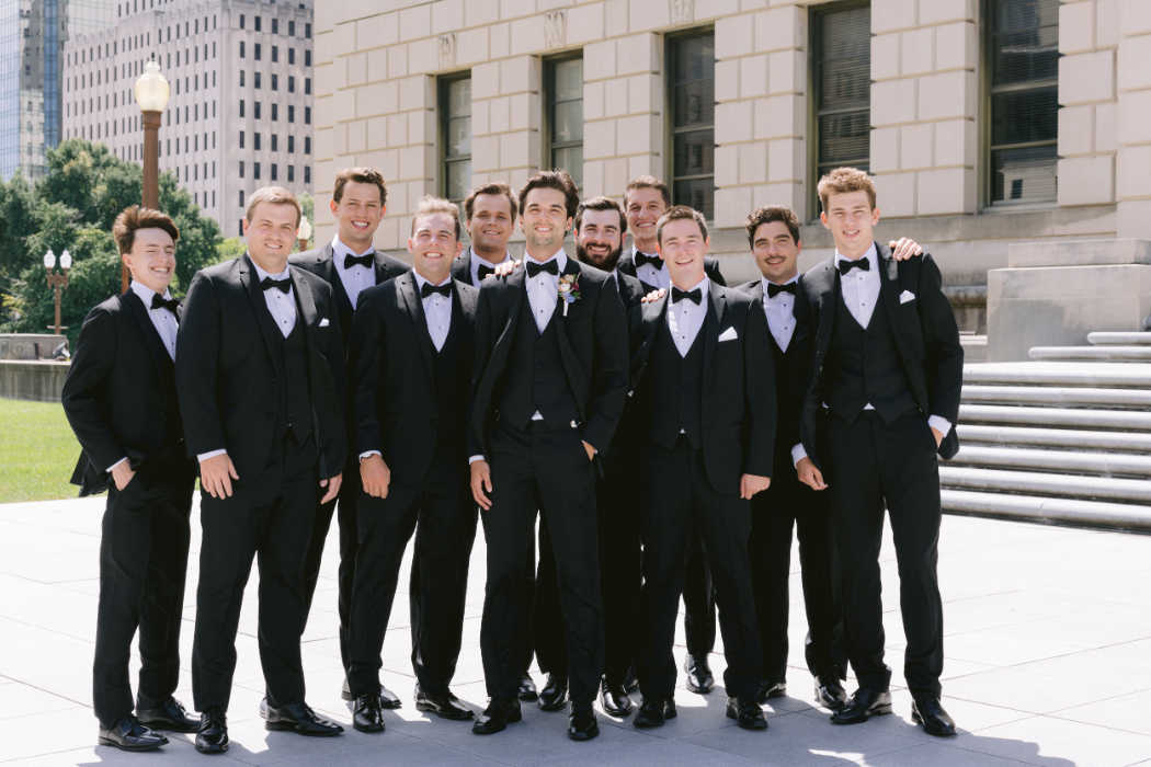 A groom stands with his groomsmen, all dressed in black tuxedos, posing in front of a grand city building. They are smiling at the camera.