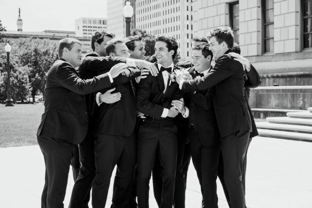 A groom in a tuxedo is surrounded by groomsmen in black suits, laughing as they embrace him in a celebratory group hug. The moment is captured outdoors in a city setting.