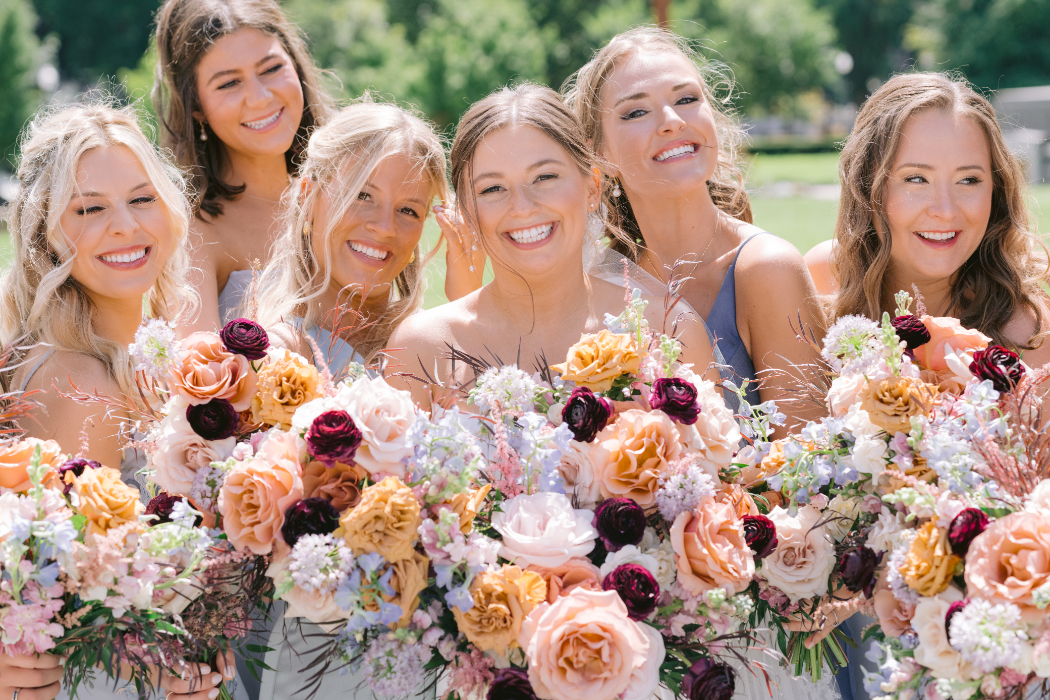 A group of bridesmaids in blue dresses stand together, holding floral bouquets while smiling. 