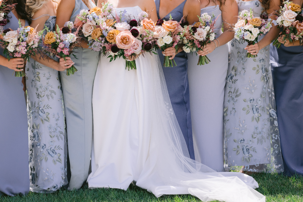 A close-up of a bride and her bridesmaids holding large bouquets of flowers in shades of pink, peach, and burgundy.