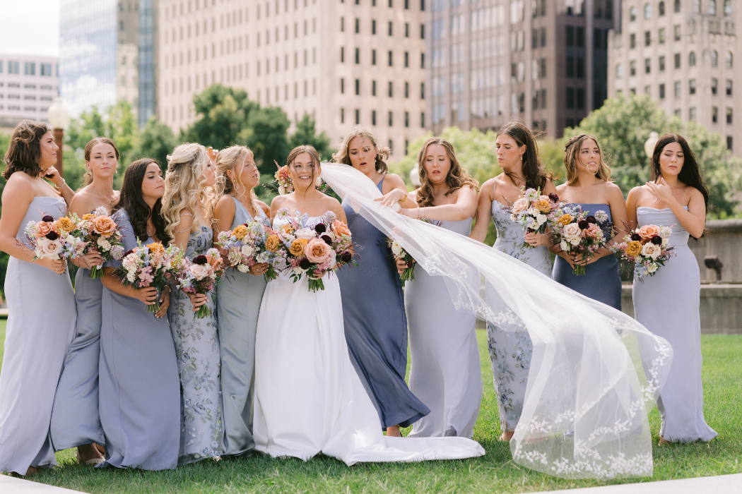 A bride and her bridesmaids holding large bouquets of flowers in shades of pink, peach, and burgundy.