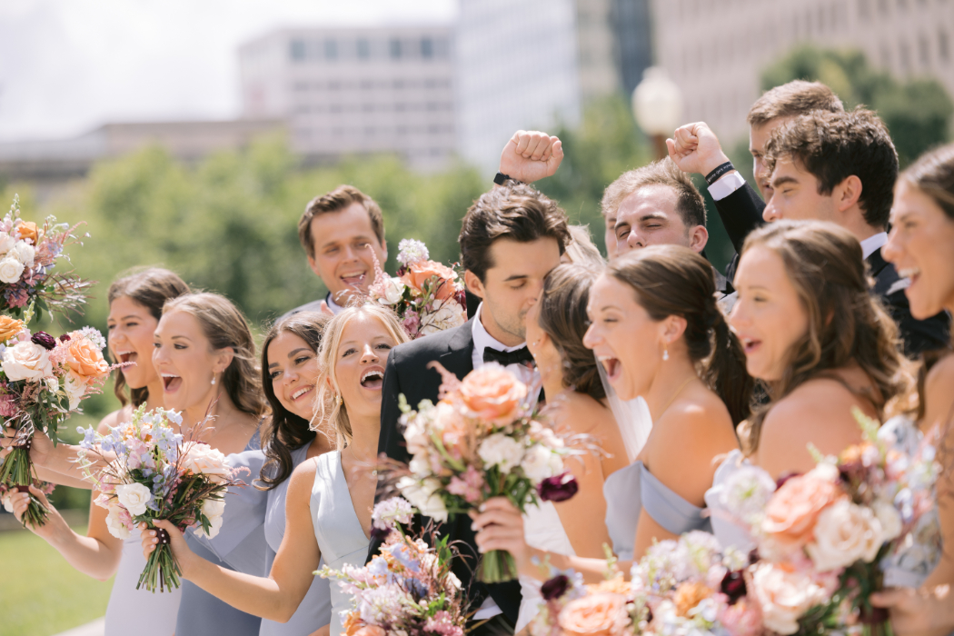 A bride and groom celebrate with their wedding party outdoors. The bridesmaids in blue dresses and groomsmen in black tuxedos cheer while holding colorful bouquets.