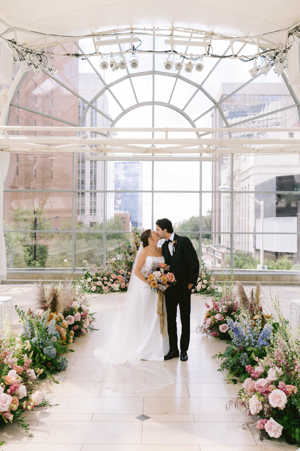 A bride and groom share a kiss in a glass-enclosed venue with city buildings visible through large arched windows. The aisle is lined with lush floral arrangements in pastel and bold colors.