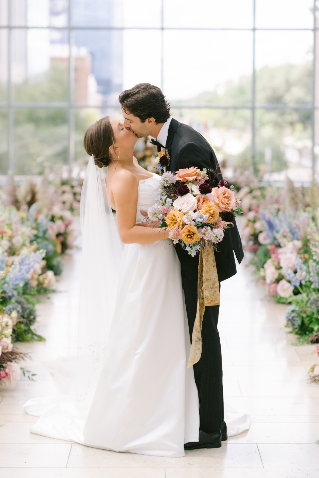 A groom in a black tuxedo kisses his bride, who holds a bouquet of pink, peach, and burgundy flowers. They stand in a bright venue with floor-to-ceiling windows and floral arrangements along the aisle.