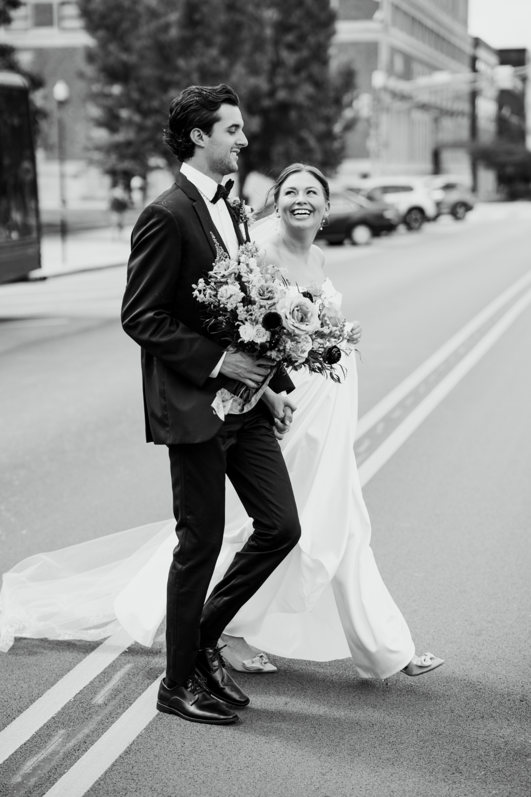 A bride and groom walk hand in hand across a city street. The groom wears a black tuxedo, and the bride wears a flowing white gown, smiling as she looks up at him while holding a bouquet.