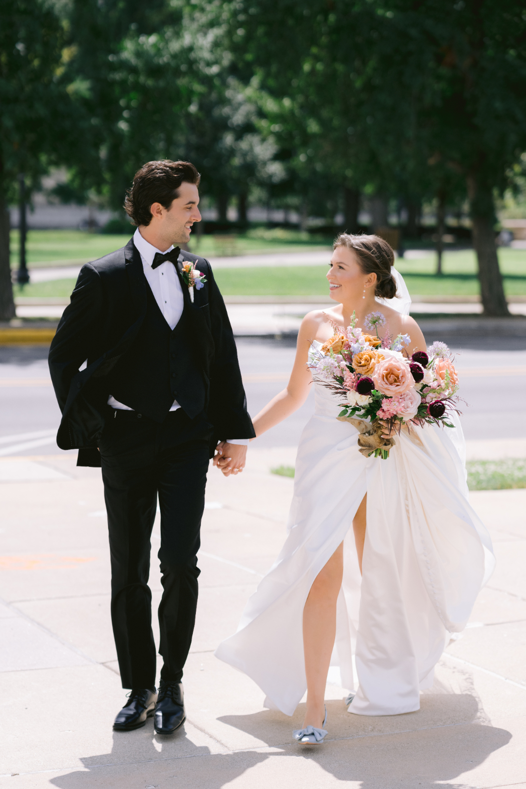 A bride and groom walk on a sidewalk, smiling at each other. The bride wears a white dress with a slit, and the groom wears a black tuxedo. She holds a bouquet of pastel and deep-colored flowers.