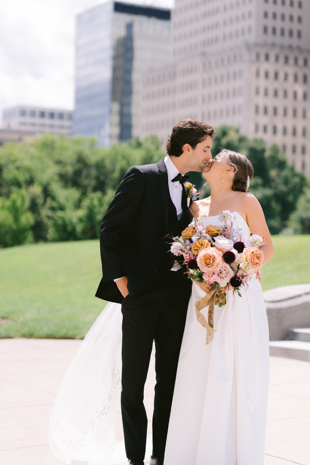 A groom in a black tuxedo kisses his bride, who holds a bouquet of pink, peach, and burgundy flowers. They stand in a park with tall city buildings in the background.