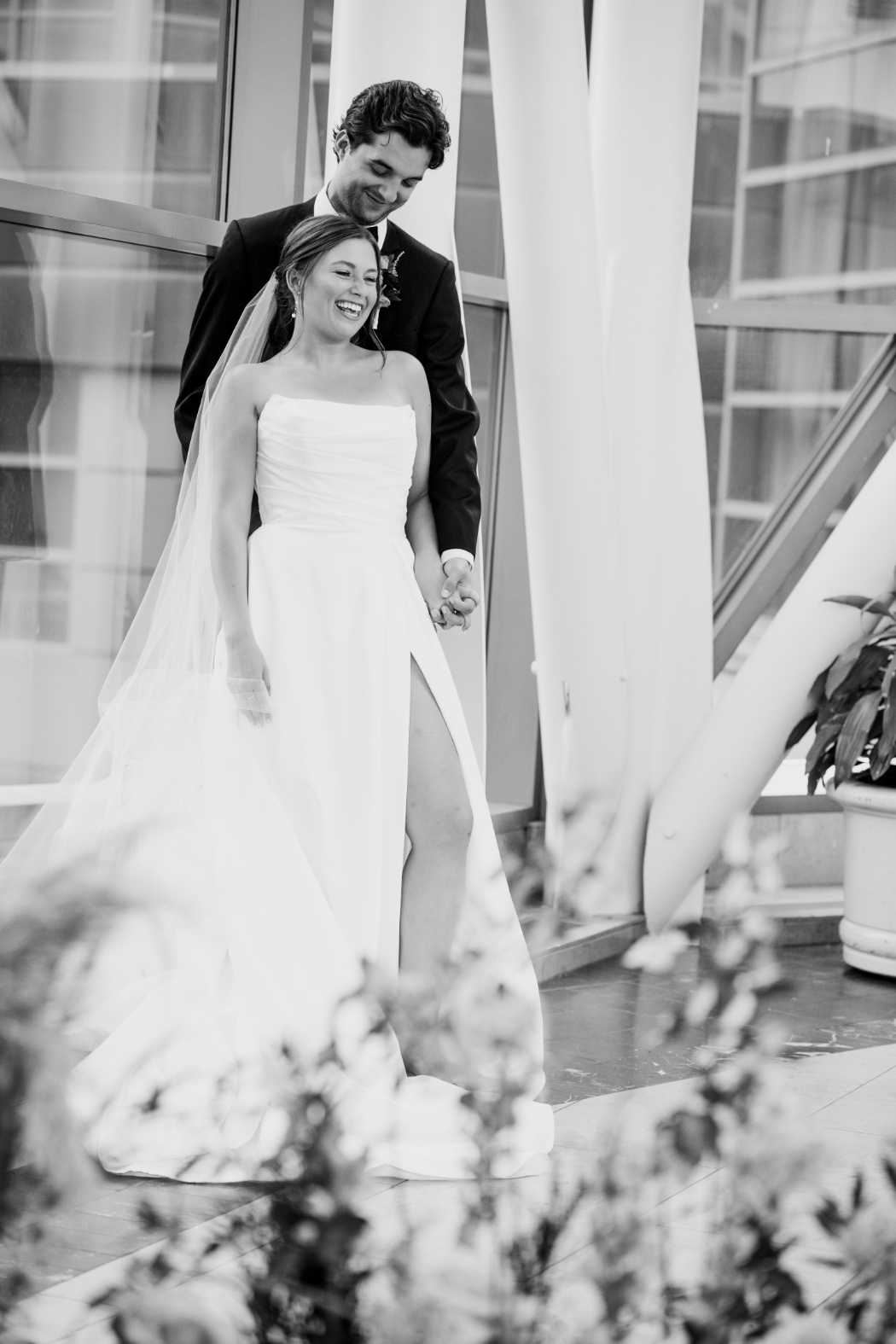 A groom stands behind his bride, holding her hand as she laughs. They are in a modern venue with large windows and structural beams, with blurred flowers in the foreground.