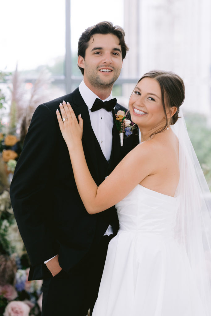 A bride and groom smile at the camera. The bride wears a strapless gown and veil, resting her hand on the groom’s chest. The groom wears a black tuxedo with a boutonniere.