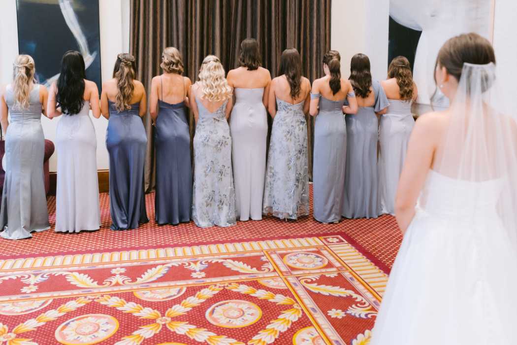 The bride's bridesmaids stand in a row, backs turned, wearing coordinated shades of blue and gray dresses, awaiting the bride’s reveal.