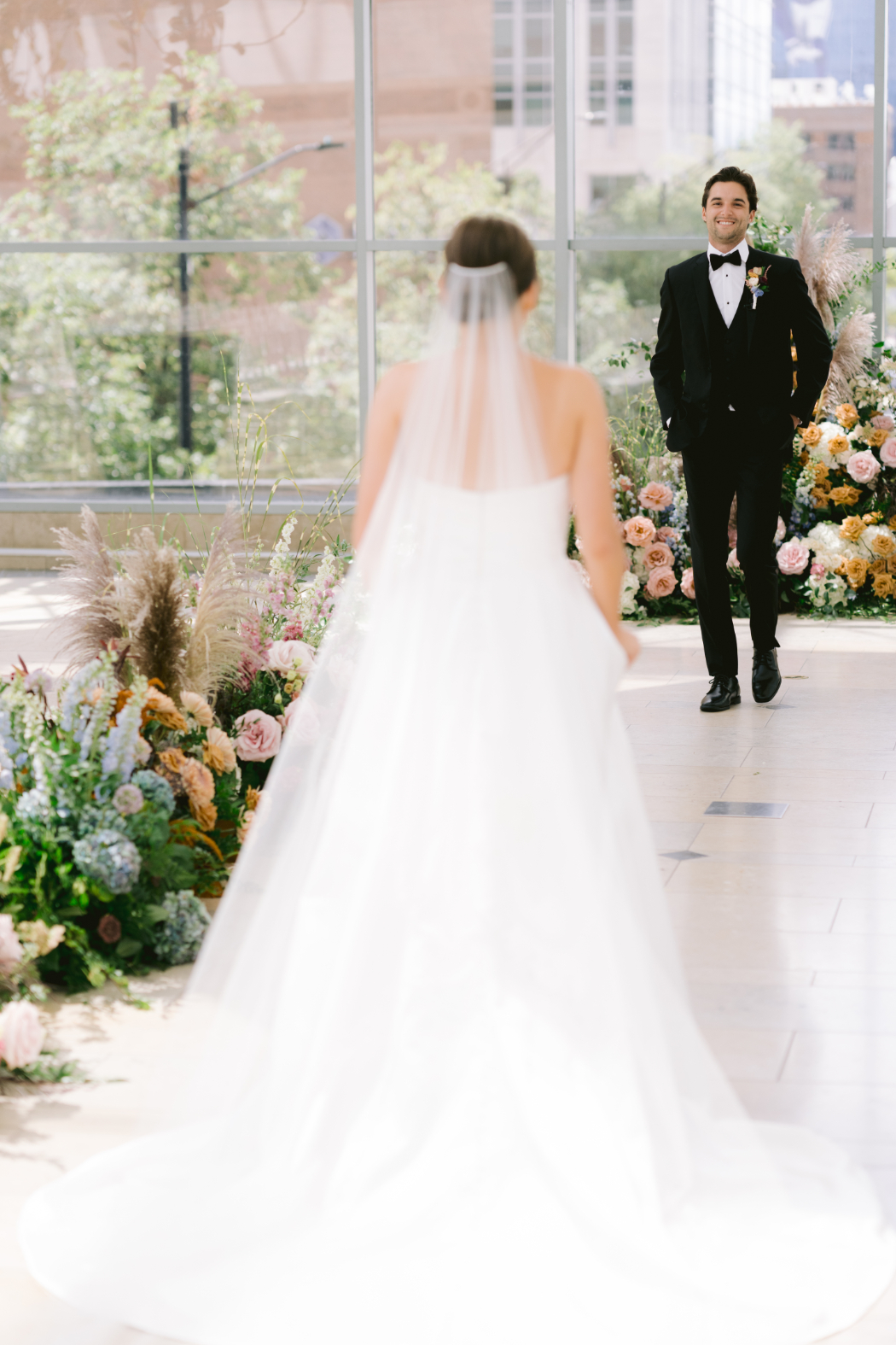 The bride, in a white gown and flowing veil, walks toward her groom for their first look, with a floral-adorned aisle leading the way.