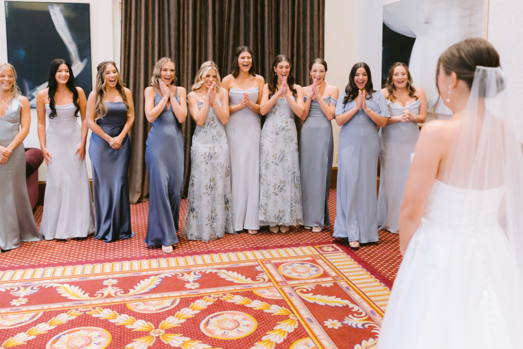 The bridesmaids react with excitement and joy, hands clasped, as they turn around to see the bride in her wedding gown.