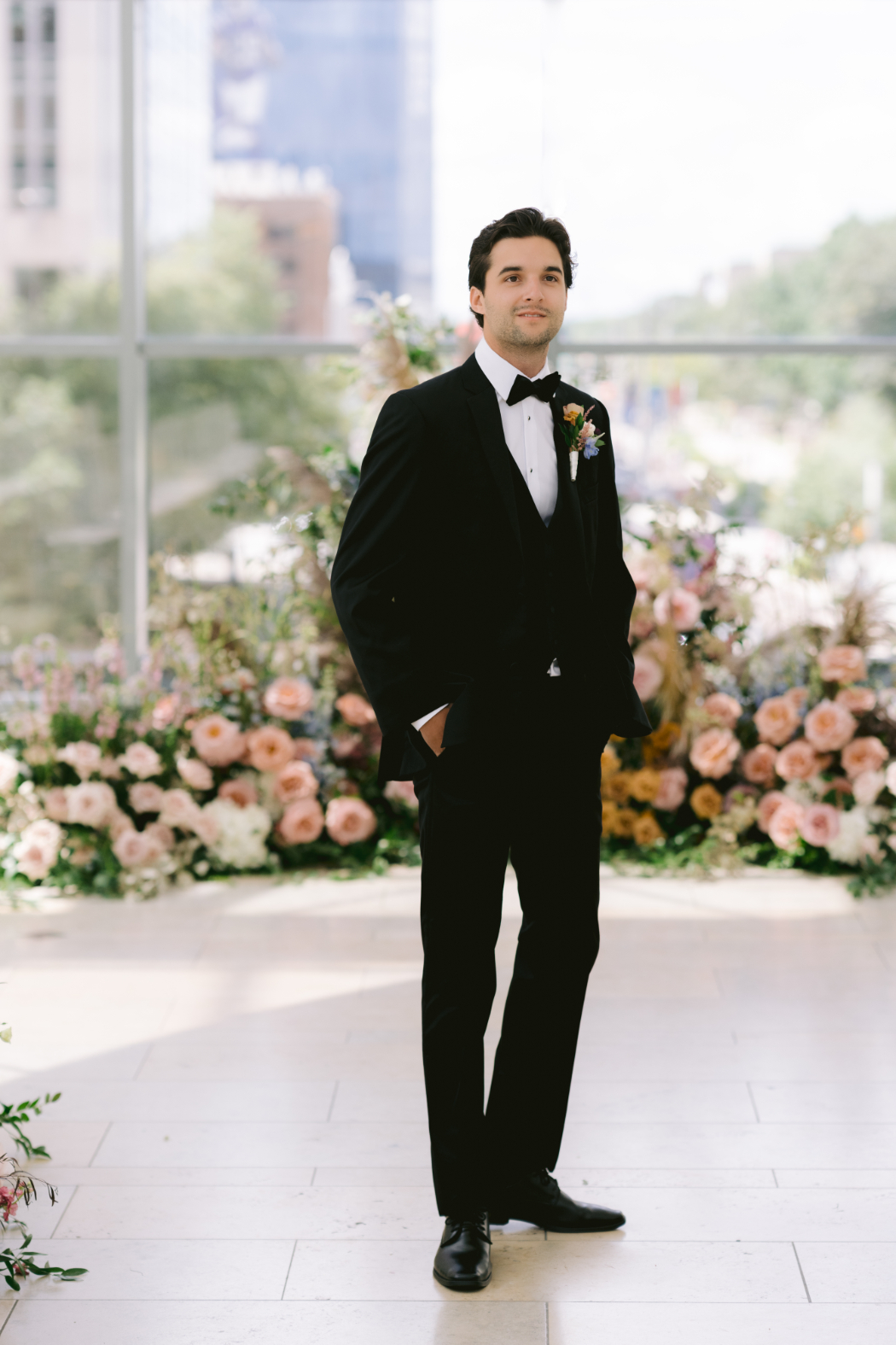 The groom stands in front of a floral-lined aisle at the Arts Garden, dressed in a black tuxedo, hands in pockets, awaiting his bride.