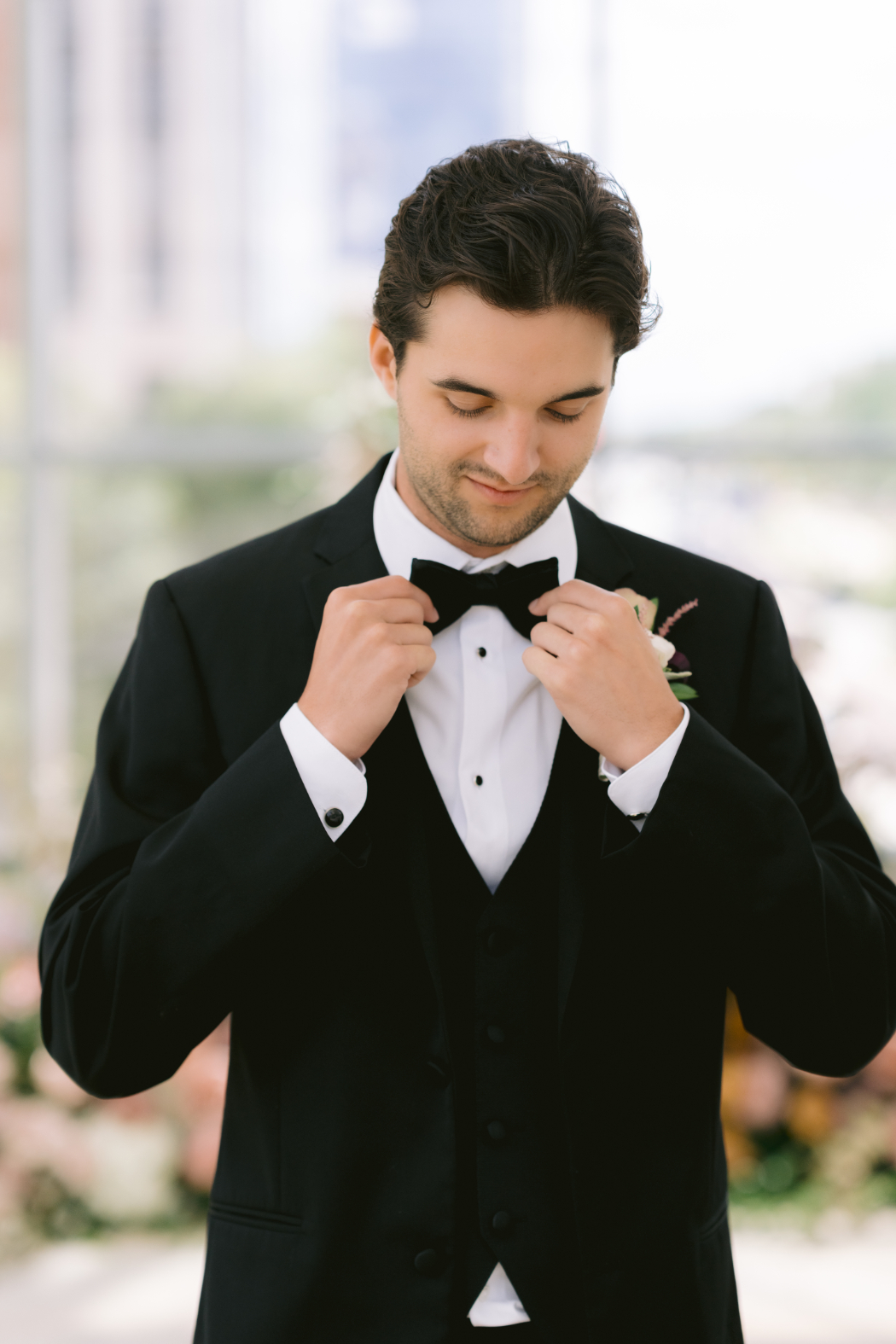 The groom adjusts his black bow tie while standing in front of a soft floral backdrop.