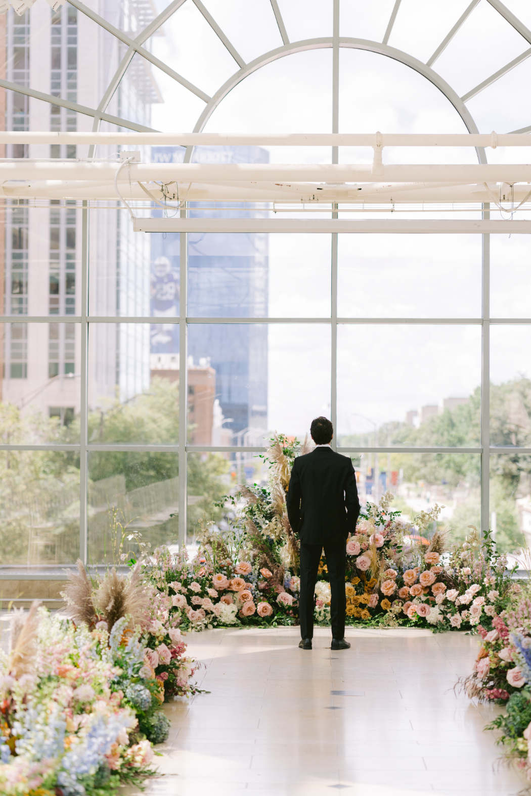 The groom stands with his back to the camera, gazing out of a large window with a cityscape view, surrounded by lush floral arrangements.