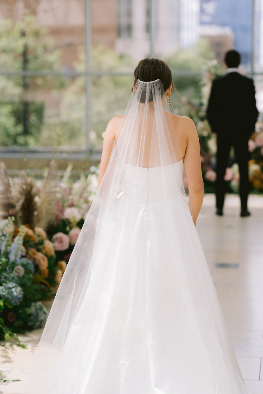 The bride, in a white gown and flowing veil, walks toward her groom for their first look, with a floral-adorned aisle leading the way.