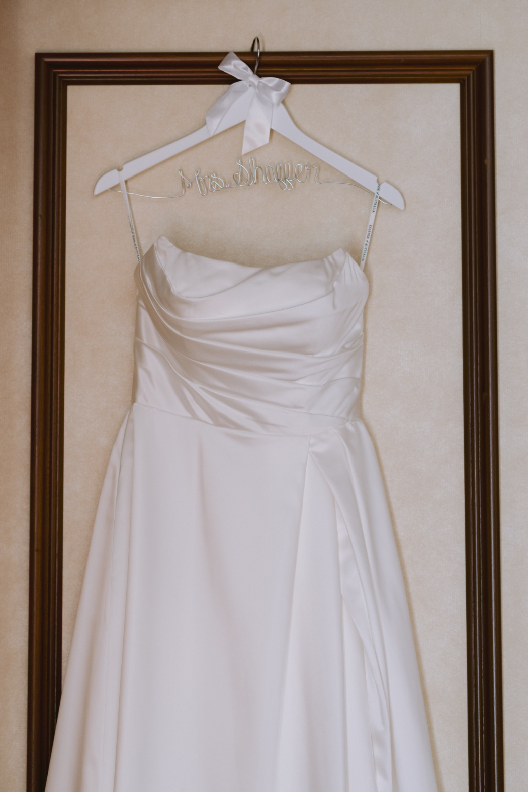 A classic white wedding gown with a ruched bodice and a flowing skirt hangs on a personalized hanger with a bow, framed elegantly.