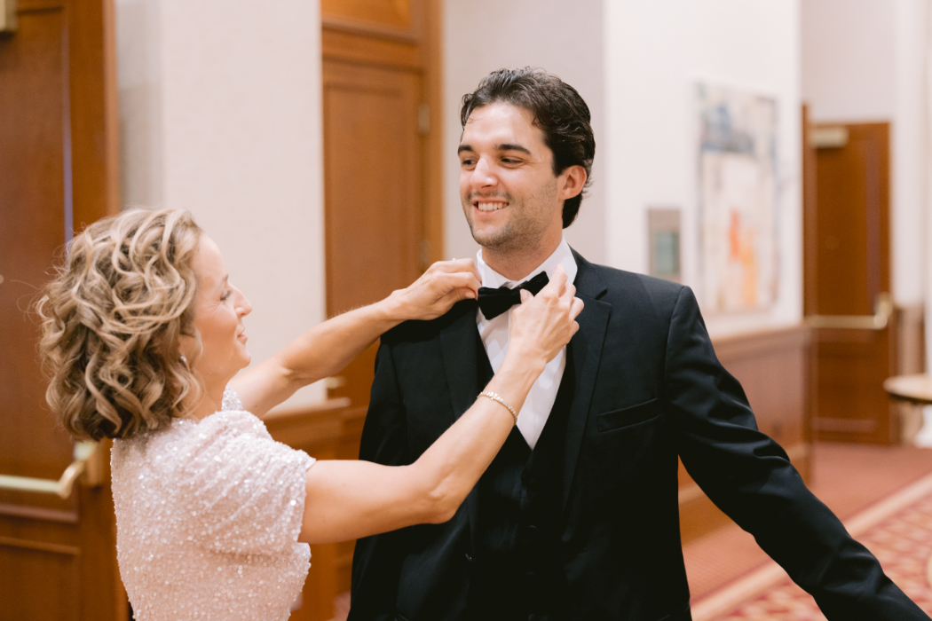 A groom's mother adjusts his bowtie as he smiles.