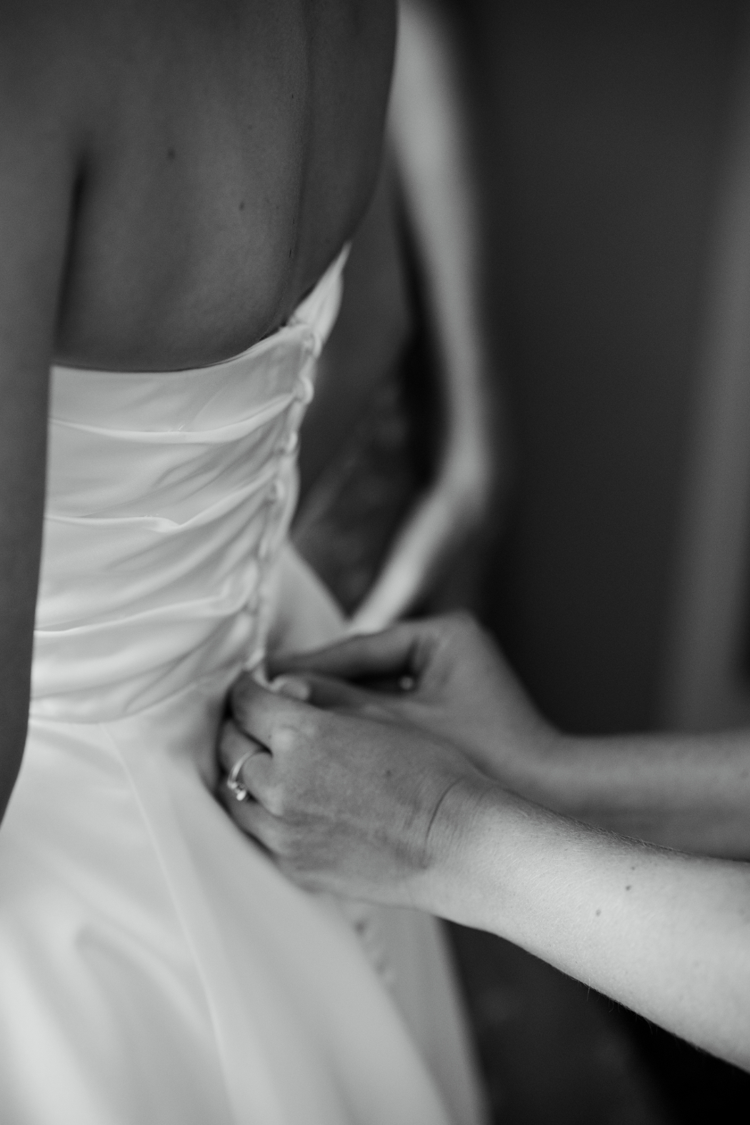 A black-and-white close-up of hands fastening the back of a bride’s dress.