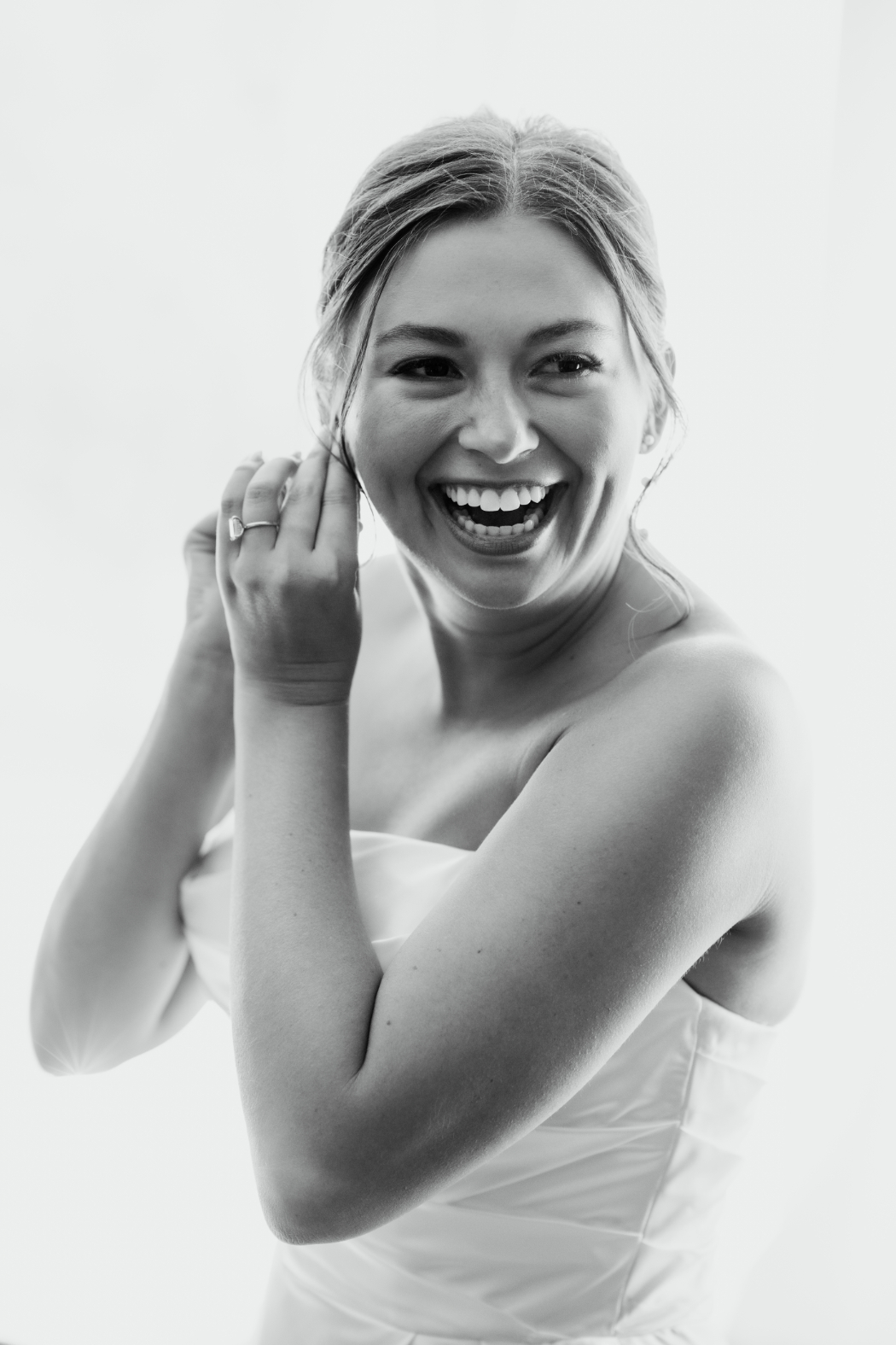 A radiant black-and-white portrait of a bride laughing as she puts on earrings.