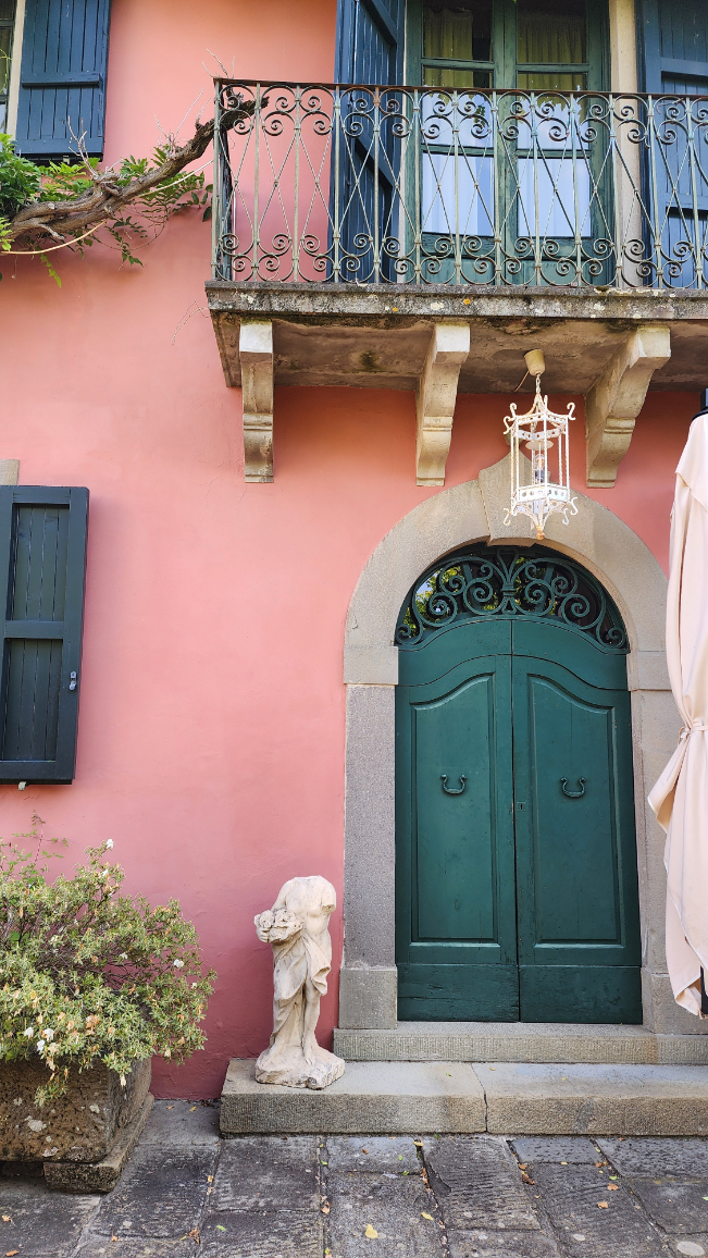 A European courtyard with a headless marble statue in front of a pink stucco wall and green shutters.