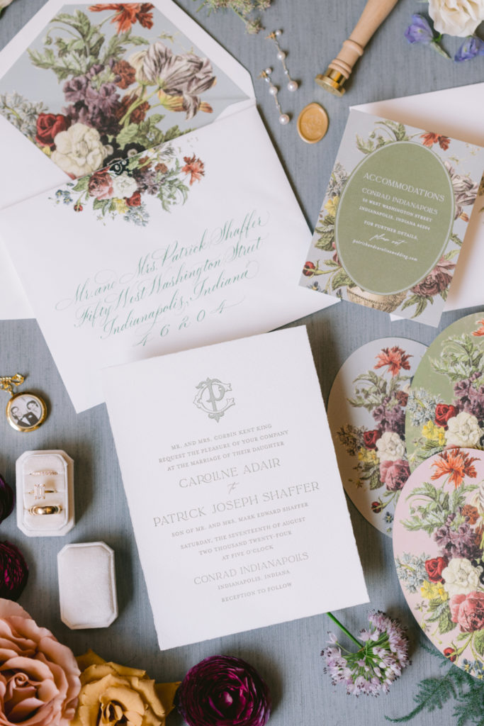 A wedding invitation suite with floral details, calligraphy envelope, gold wax seal, and accessories, displayed on a soft blue fabric background.