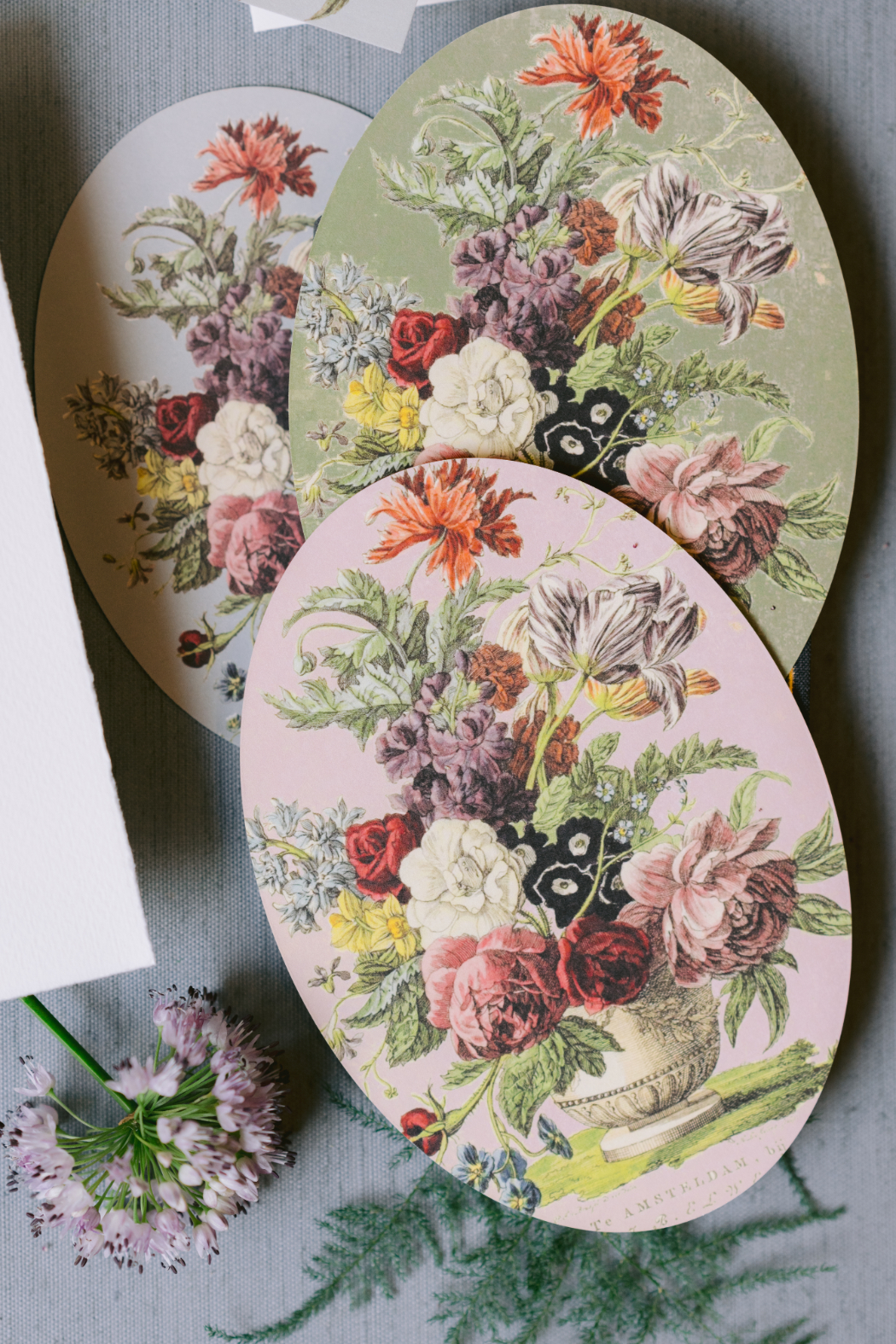 Three oval-shaped wedding stationery pieces with floral still-life designs in green, pink, and blue, inspired by Dutch Masters paintings.