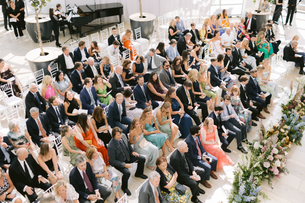 Wedding guests sit on white chairs at arts garden venue, waiting for the ceremony to begin. Some guests hold programs, and musicians play a piano in the background.