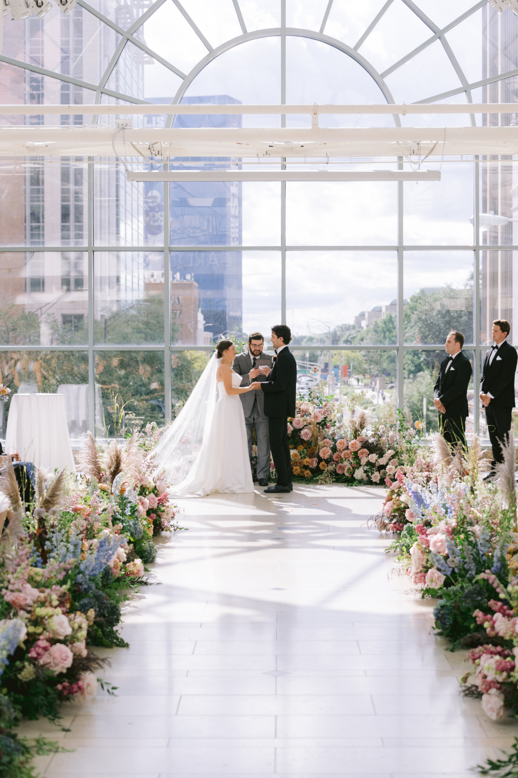 Bride and groom at the altar, surrounded by floral arrangements in a glass-ceiling venue with city views.