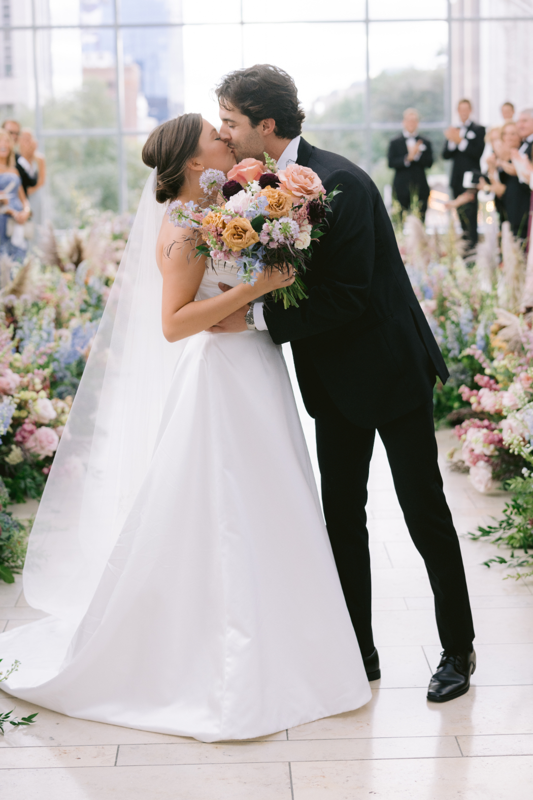 Close-up of the bride and groom kissing, holding a bouquet of peach, pink, and burgundy flowers.