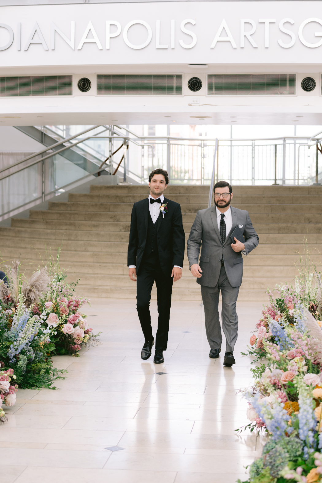 A groom in a black tuxedo walks down the aisle with an officiant at the Indianapolis Artsgarden. The aisle is decorated with colorful floral arrangements.