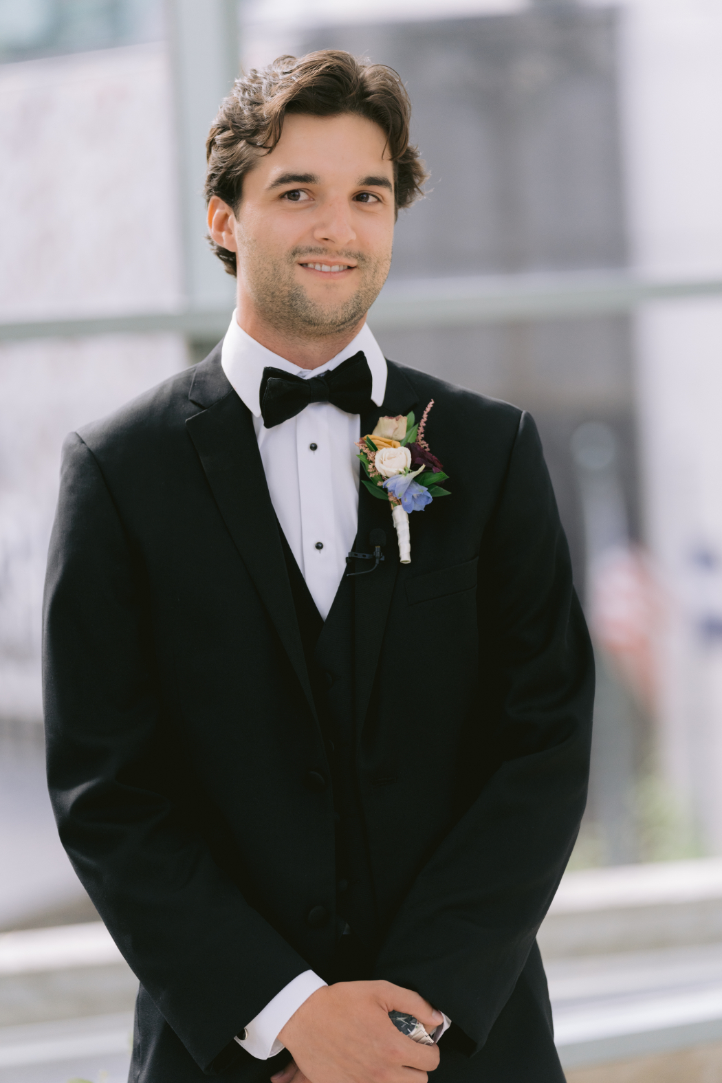 A groom in a black tuxedo stands in a bright venue, smiling slightly. He wears a boutonniere with pastel flowers and a microphone attached to his jacket.