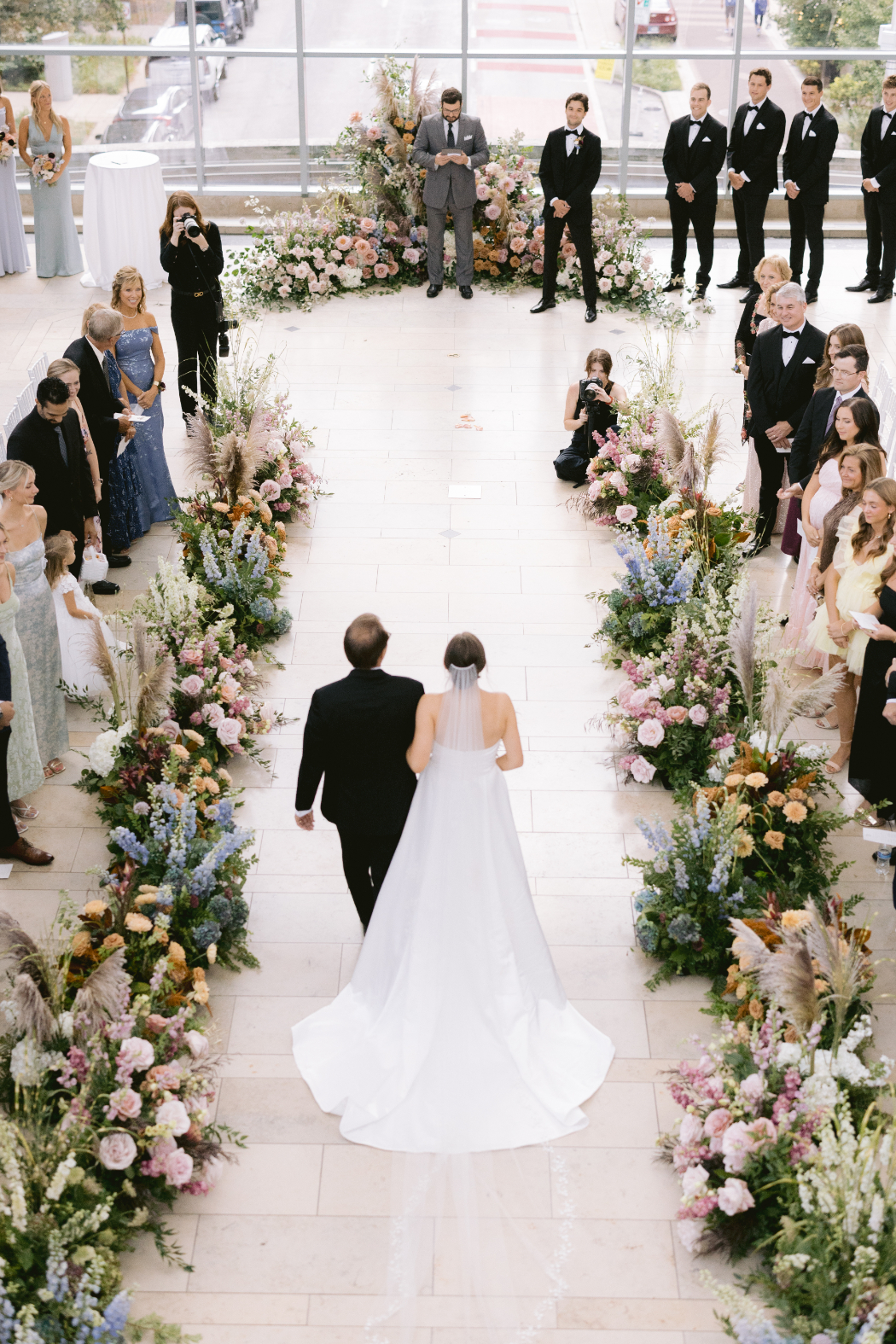 A bride walks down a flower-lined aisle with her father at a wedding ceremony in a modern venue with large windows. Guests watch from their seats.