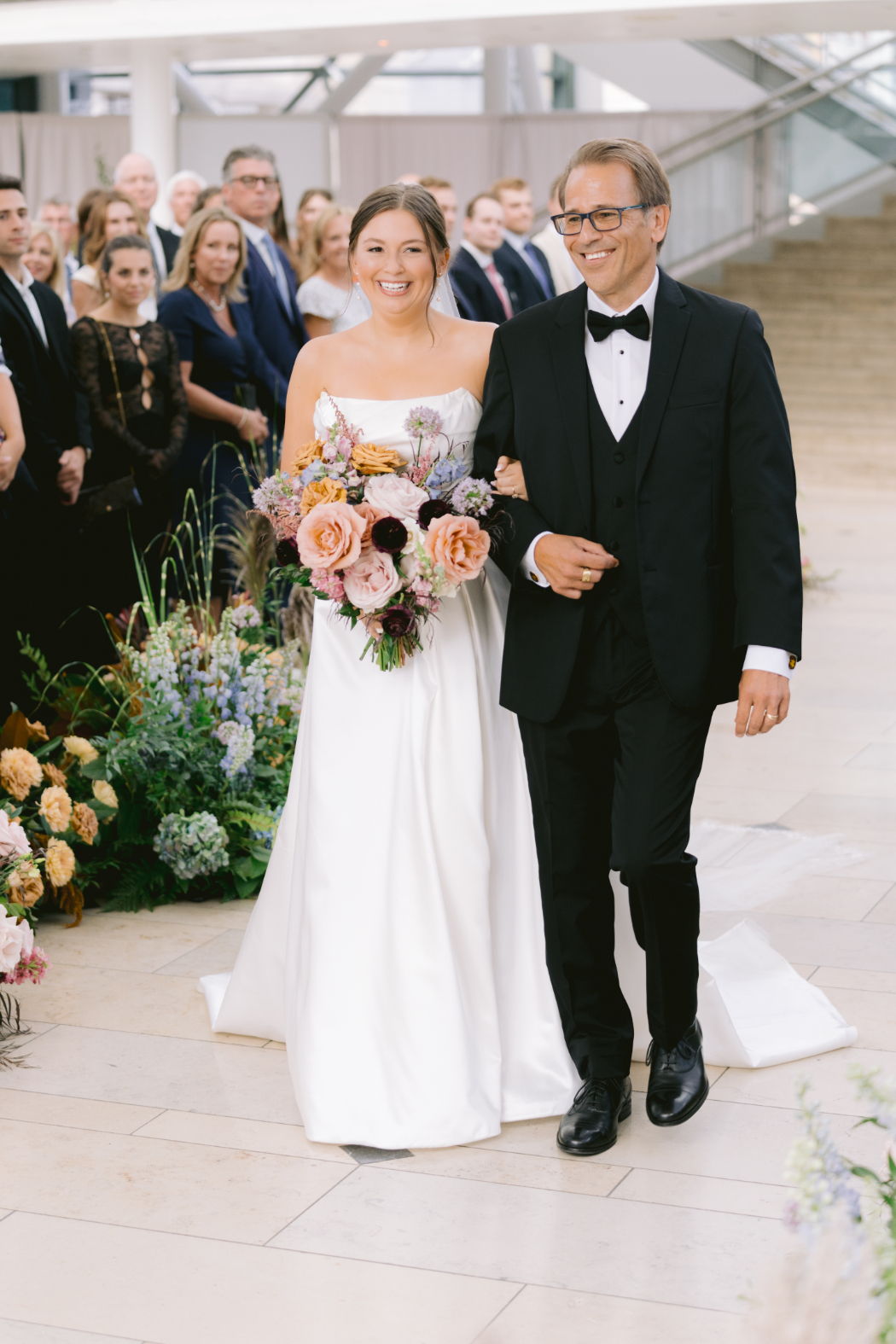 A bride, smiling, walks down the aisle with her father at her wedding ceremony. She carries a bouquet of pink, peach, and burgundy flowers, and guests watch in the background.