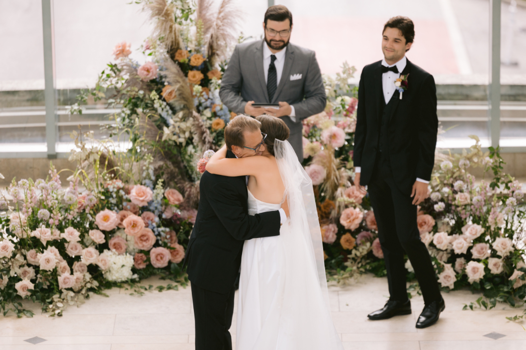 A bride hugs her father at the altar as the groom watches, smiling. The ceremony is set against a floral backdrop with an officiant reading from a book.