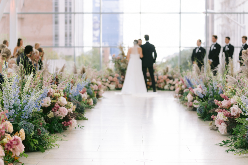 A bride and groom stand at the altar in a modern glass venue with large floral arrangements along the aisle. Groomsmen in black tuxedos stand in the background.