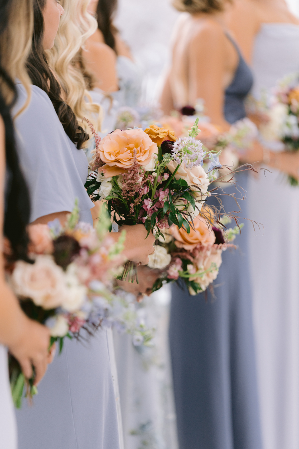 Bridesmaids in blue dresses hold bouquets of pastel and deep-colored flowers during the wedding ceremony. The image focuses on the floral arrangements in their hands.