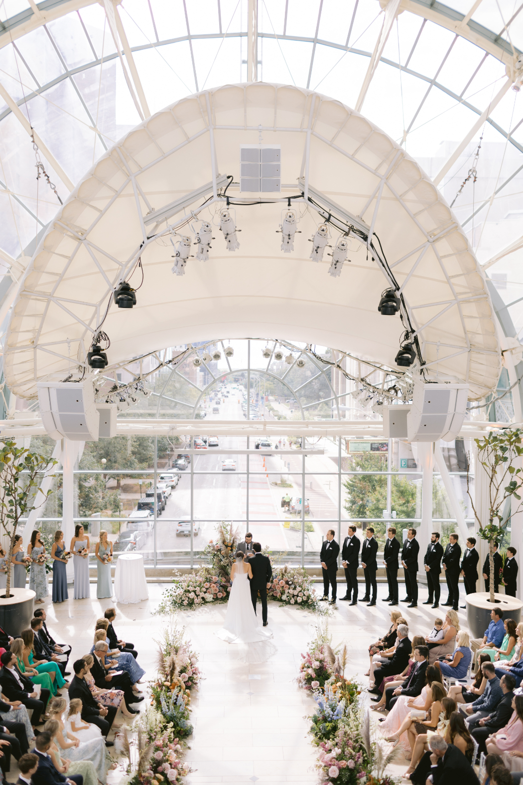 Bride and groom at the altar, surrounded by floral arrangements in a glass-ceiling venue with city views.